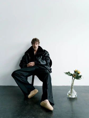 Stylish man in oversized jacket poses indoors while seated. Features sunflower arrangement, dark monochromatic tones, and studio lighting.