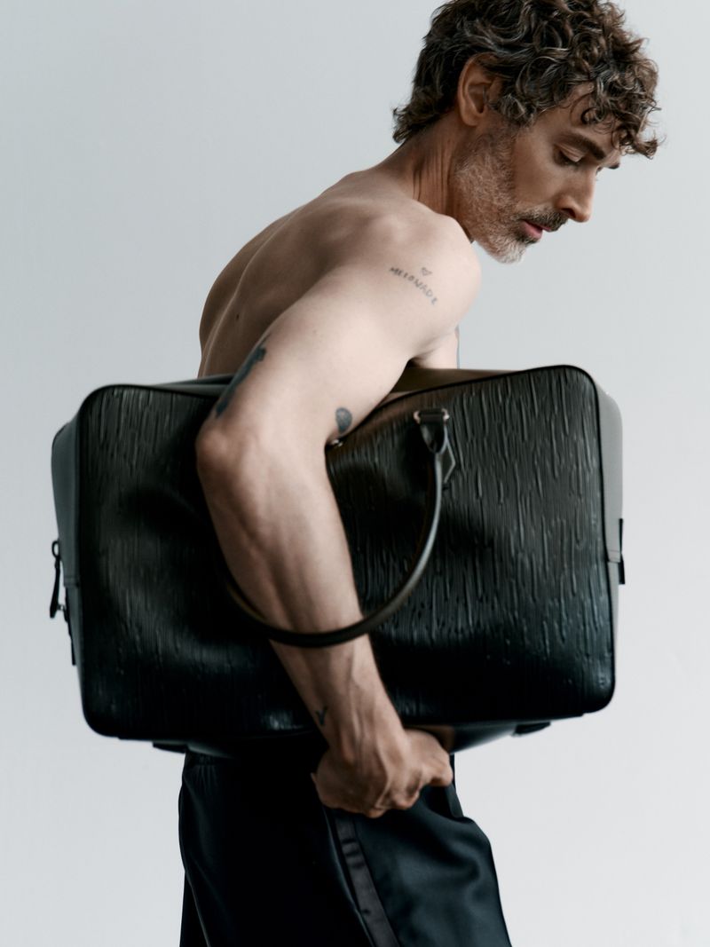 A shirtless man with curly hair carries a textured black duffel bag in front of a plain background, showcasing a minimalist fashion aesthetic.