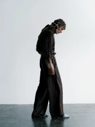 Richard Biedul posing in profile wearing a textured brown knit sweater and wide-leg brown trousers against a white studio wall.