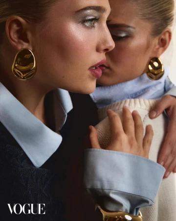 Close-up of two blonde women in blue and black collared shirts with gold earrings and bracelets in a Vogue editorial fashion shot.