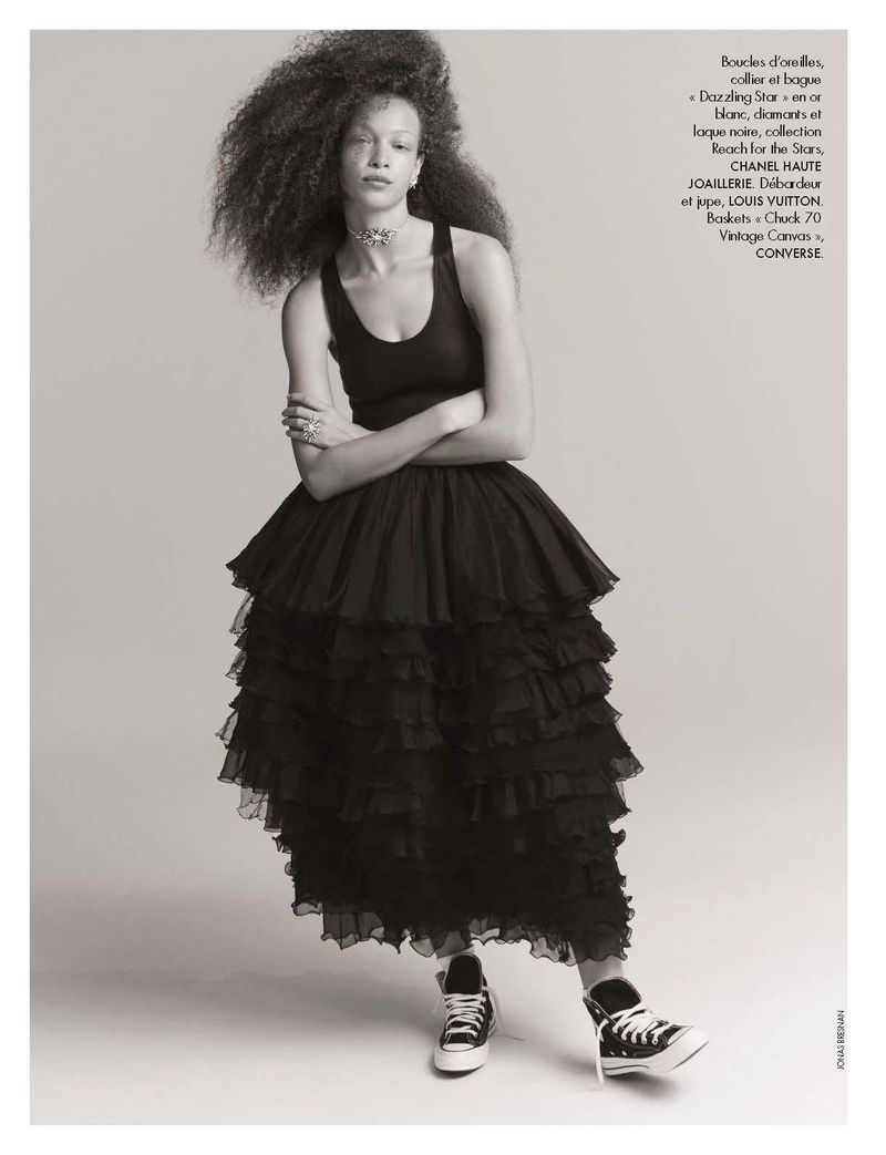 Black and white portrait of a woman with voluminous dark hair wearing a black dress with layers and black and white sneakers.