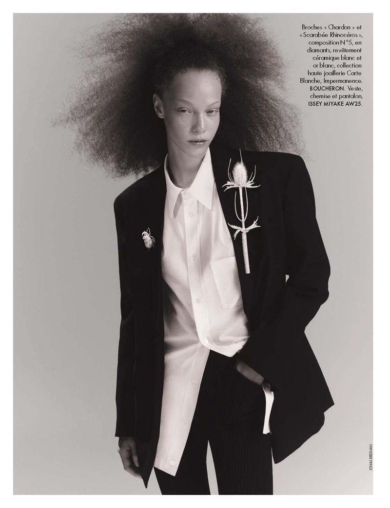 Fashion editorial shot of a Black model with afro hair wearing a black blazer with floral brooches, white blouse, and black pants.
