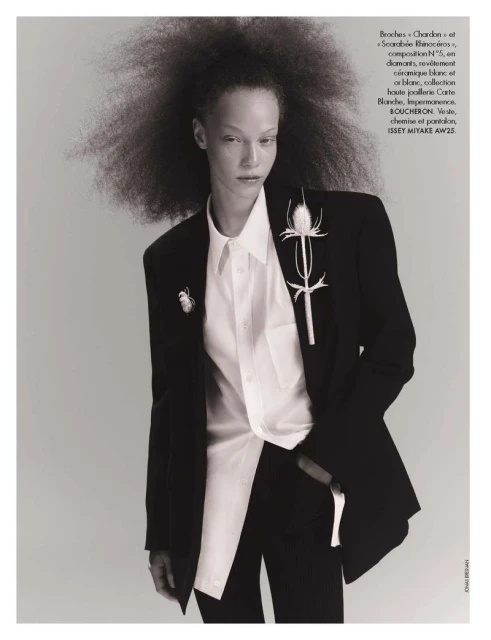 Model with voluminous afro hairstyle wearing a black blazer and white collared shirt against grey seamless background.