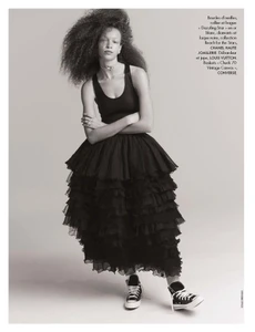 Fashion model posing with arms crossed in a black tank top, tiered ruffled black skirt, and canvas high-top sneakers on a studio backdrop.