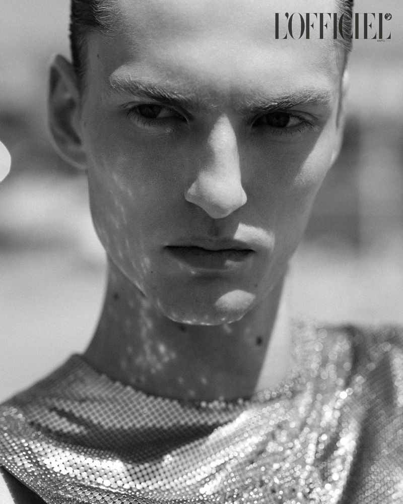 Portrait of a fair-skinned adult male with short hair wearing a shiny metallic top, with an outdoor background and natural lighting.