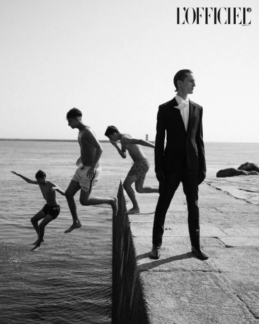 Black and white fashion shot featuring a male model in a suit standing on a pier, with three young men jumping into the ocean.