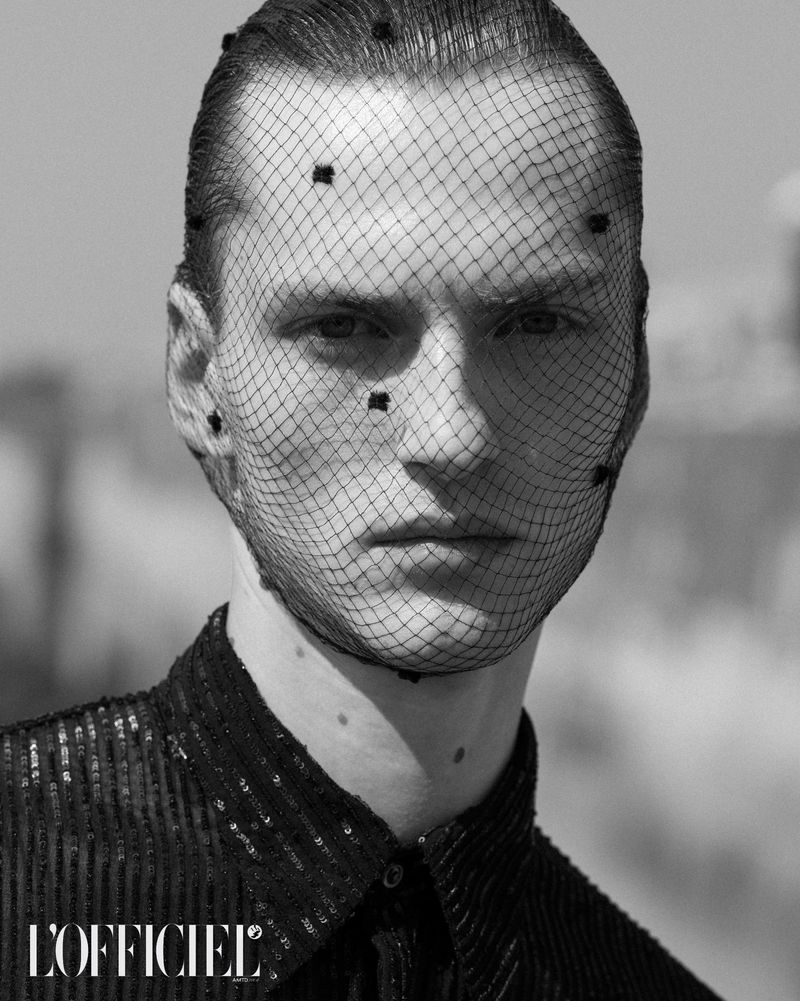 Monochrome portrait of a man with a net veil over his head, wearing a sequined top, and shot at eye-level outdoors during the day.