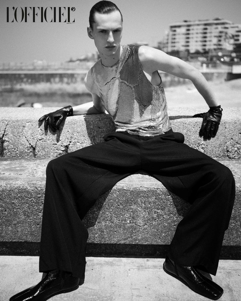 Black and white fashion portrait of a thin male model sitting on concrete with a water view in the background, wearing gloves and a shiny vest.