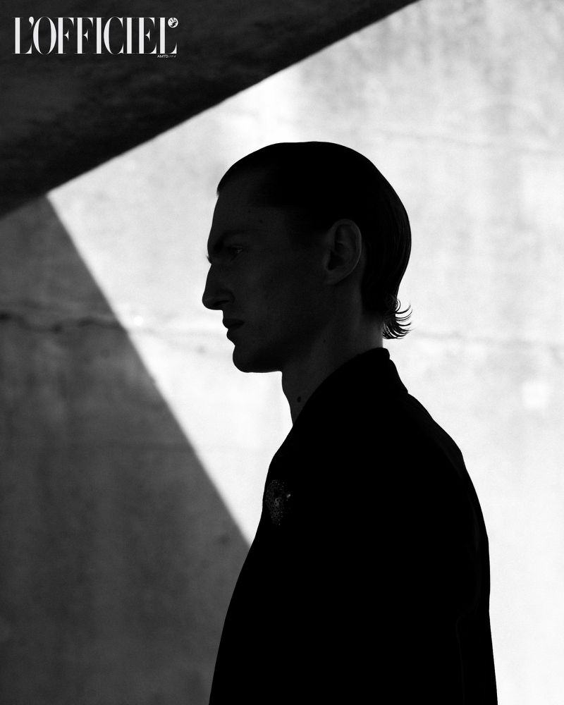 Black and white studio shot of a man in profile, showcasing sharp facial features, dark jacket, and the L'Officiel magazine logo.