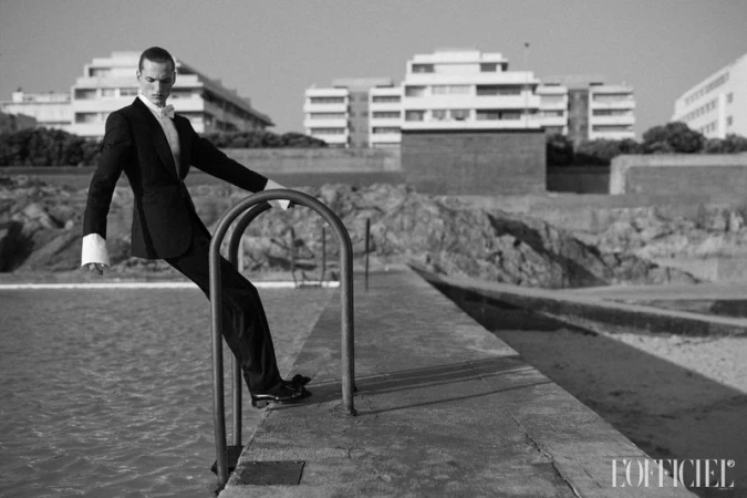 Black and white fashion shot of a man in a tuxedo leaning on a pool ladder, with modern buildings in the background on a sunny day.