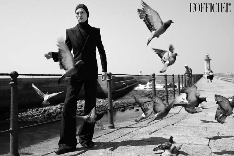 Monochrome fashion shot of a male model in a suit surrounded by pigeons on a pier, with a lighthouse in the background under bright daylight.