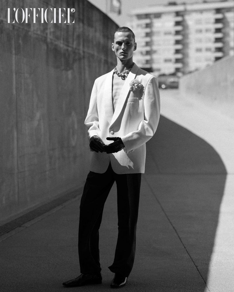 Monochrome fashion shot of a slender adult male model in an off-white blazer, black gloves and pants, and a statement necklace, posing outdoors.