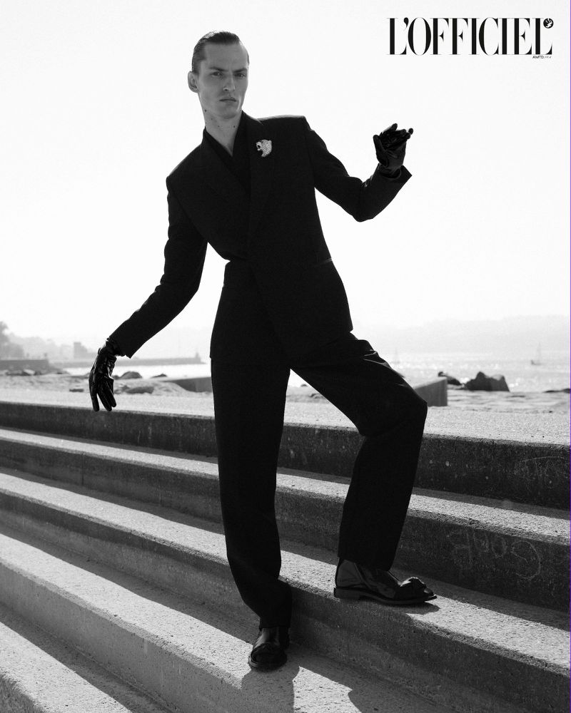 Fashion editorial shot of a slender adult male wearing a black suit and gloves, posing on concrete stairs with ocean and coastline background.