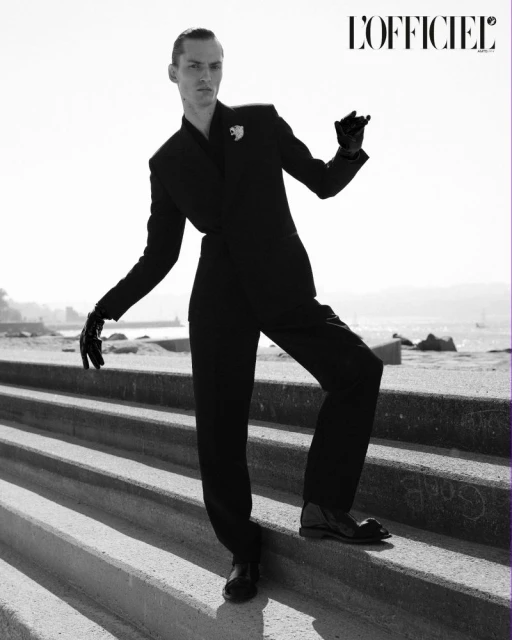 Fashion editorial shot of a slender adult male wearing a black suit and gloves, posing on concrete stairs with ocean and coastline background.