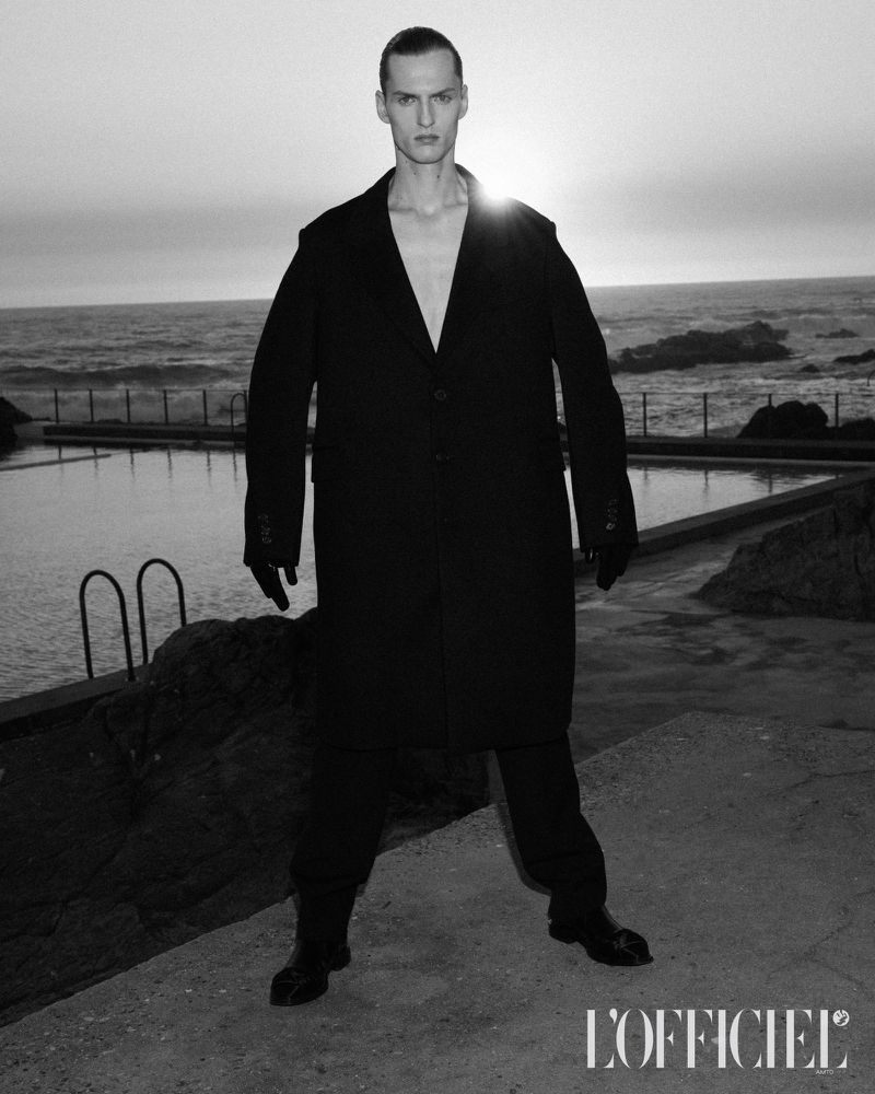 Black and white fashion shot of a man in a coat standing near an ocean pool at sunset, wearing dark clothing and gloves with the L'Officiel logo.