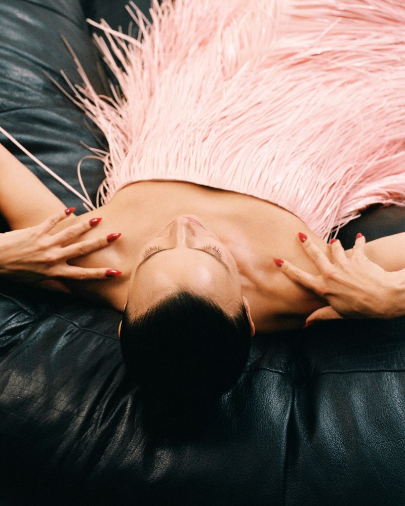 Adult woman lays on black leather couch wearing a pink fringed dress with hands resting on chest. Shot from directly above.