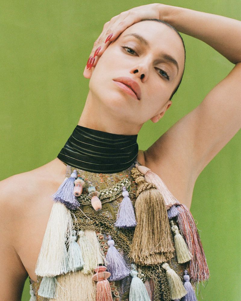 Close-up of a fair-skinned woman with red nails, wearing a black choker and a sleeveless top with multi-colored tassels against green background.
