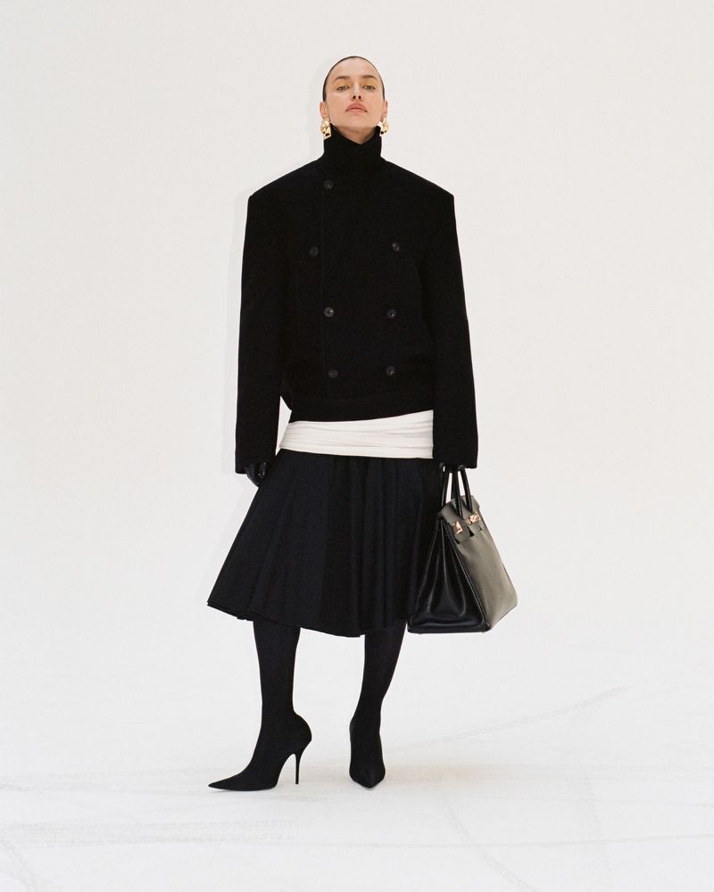 Irina Shayk posing in a high-neck black jacket, pleated skirt, black tights, and pointed boots against a white background.