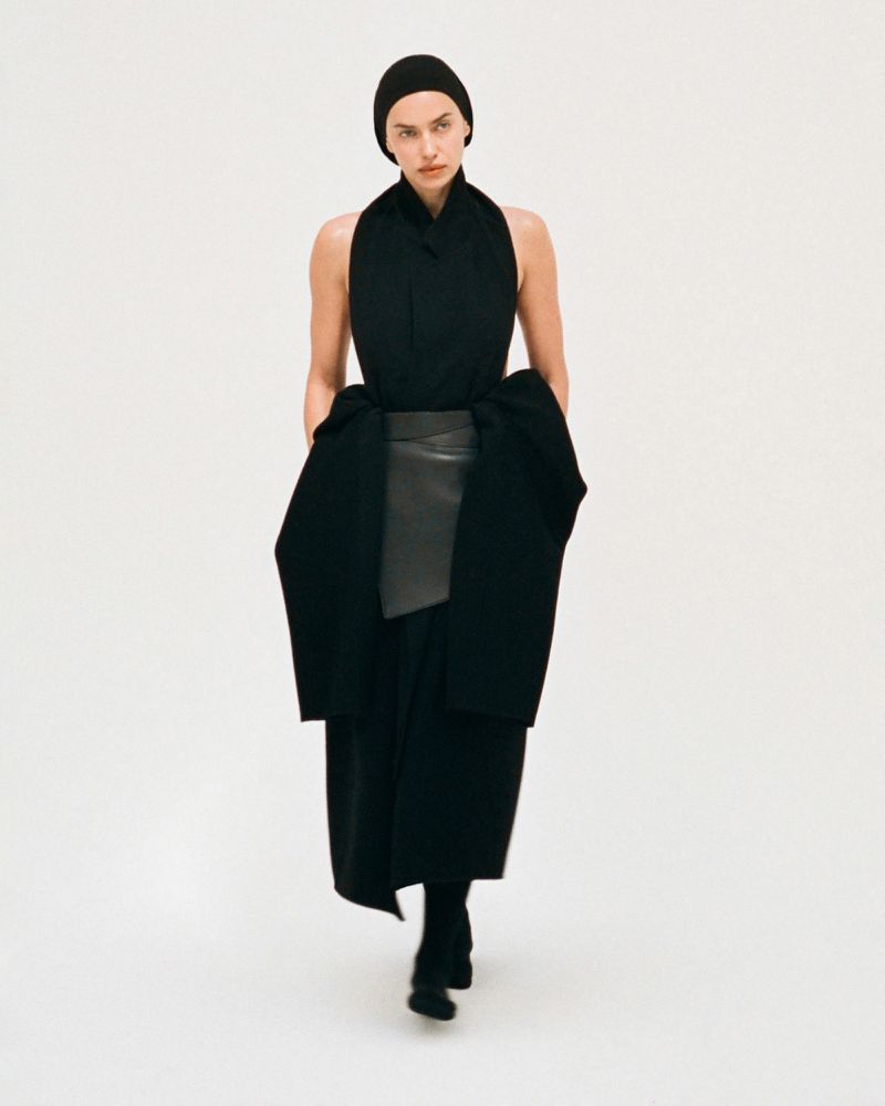 Fashionable adult female model in a black knit cap, dress, and booties standing against an off-white studio backdrop with hands in the pockets.