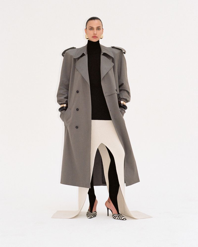 Full-length shot of an adult woman posing in a grey trench coat, black turtleneck, and patterned shoes against a white backdrop.