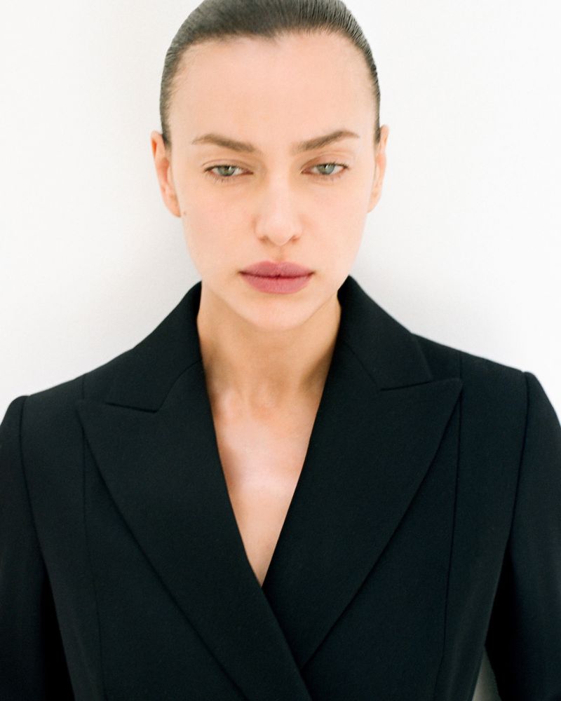 Close-up portrait of a woman with slicked-back hair wearing a black double-breasted blazer against a white background in a photo studio.