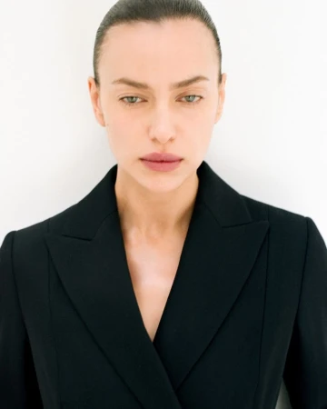 Close-up portrait of a woman with slicked-back hair wearing a black double-breasted blazer against a white background in a photo studio.