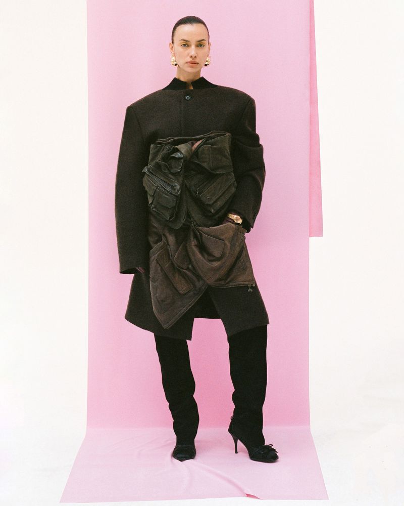 A solo fashion model poses against a pink backdrop, wearing a brown coat with cargo pockets and black heeled boots in a studio shot.