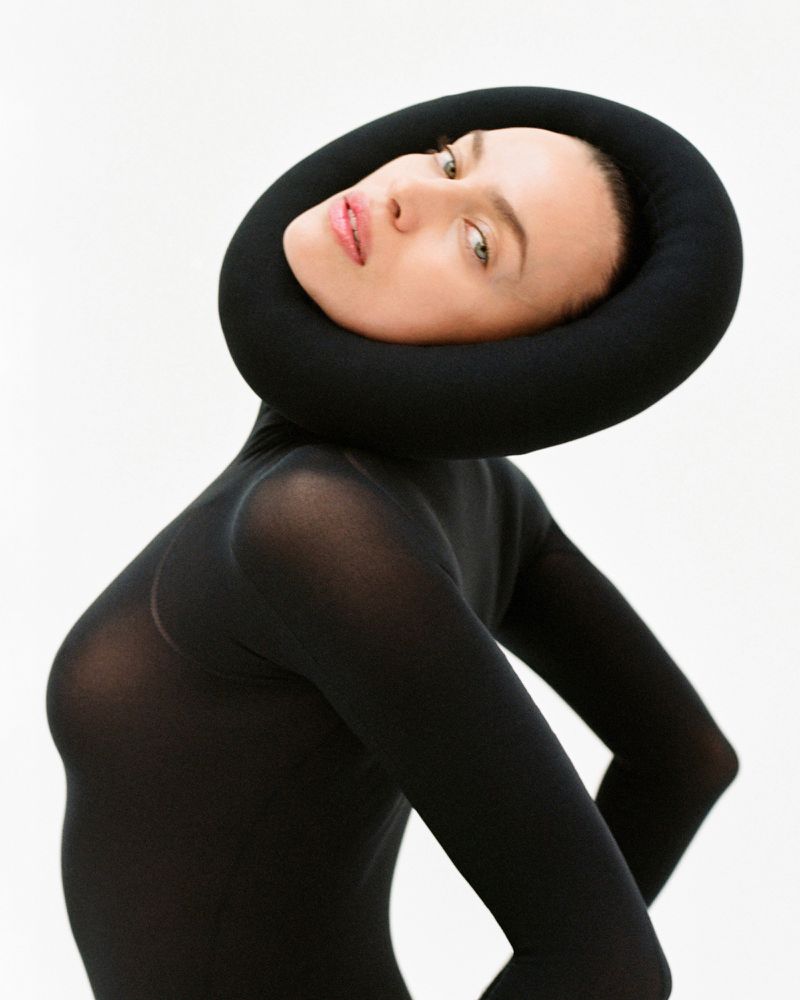 A woman in a black bodysuit and a round black headdress poses against a white background, face tilted, in a fashion-forward studio portrait.