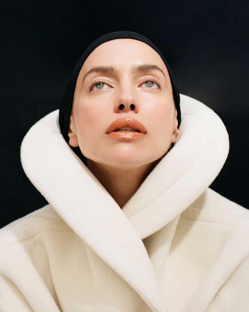 Close-up portrait of a woman with slicked-back hair wearing a white collared coat looking upward against a dark backdrop.