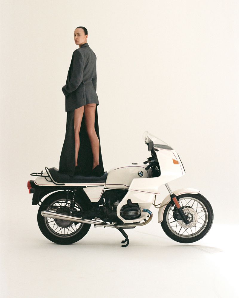 A woman stands on a white motorcycle wearing a grey suit jacket and black shoes in a white photo studio setting.