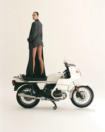A woman stands on a white motorcycle wearing a grey suit jacket and black shoes in a white photo studio setting.