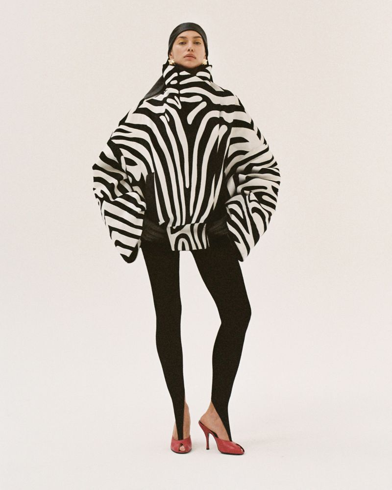 A woman in a zebra-striped sweater and leggings stands in a photo studio. The image features a fashion-forward, eye-level studio shot.