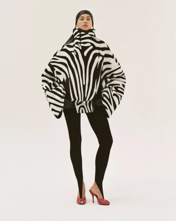 A woman in a zebra-striped sweater and leggings stands in a photo studio. The image features a fashion-forward, eye-level studio shot.
