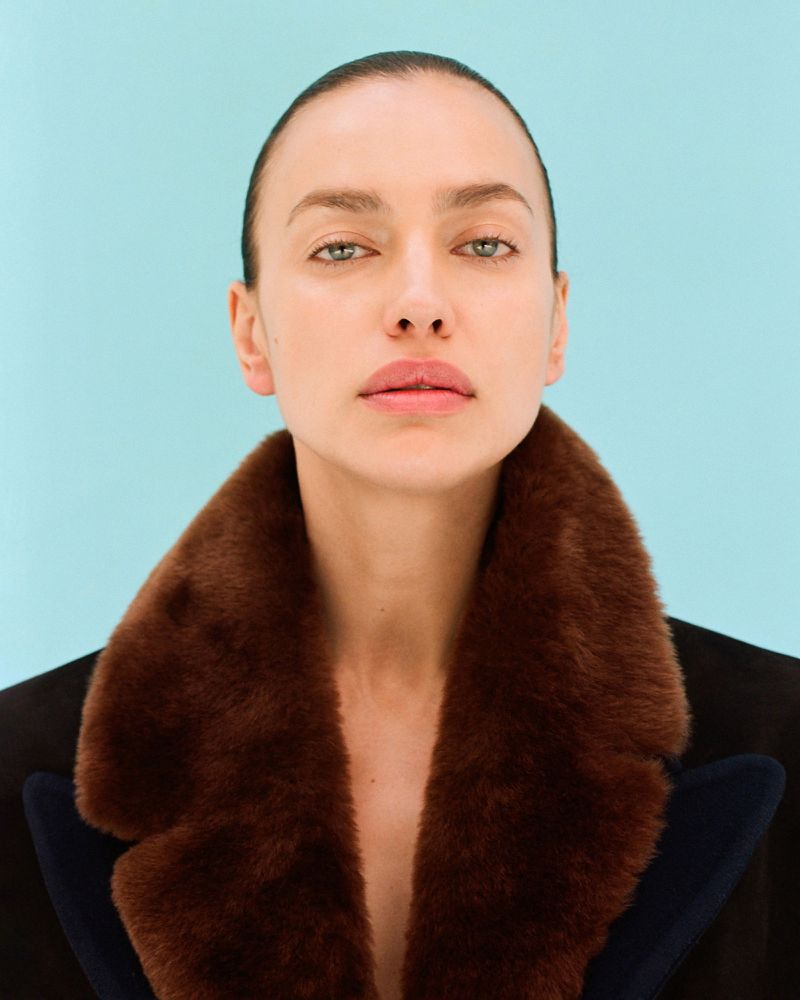 Close-up portrait of model Irina Shayk wearing a dark coat with a brown faux fur collar against a solid light blue background.