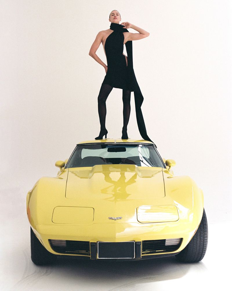Model Irina Shayk standing atop the roof of a vintage yellow sports car in a black mini dress against a white studio background.