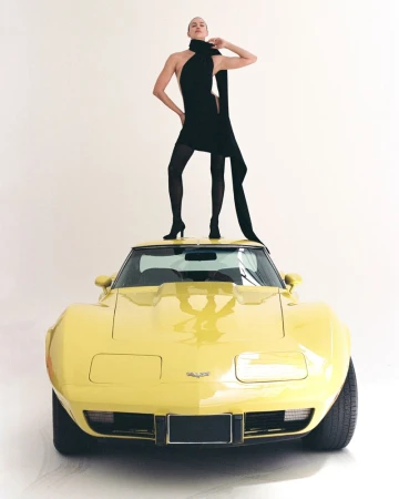 Model Irina Shayk standing atop the roof of a vintage yellow sports car in a black mini dress against a white studio background.
