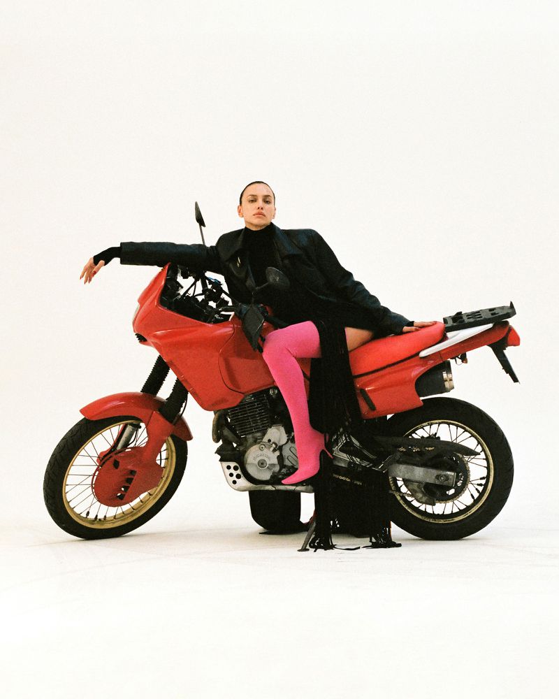 A woman in a black outfit and pink tights reclines on a red motorcycle in a studio setting against a white backdrop.