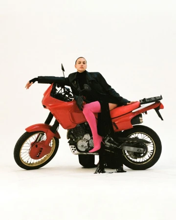 A woman in a black outfit and pink tights reclines on a red motorcycle in a studio setting against a white backdrop.