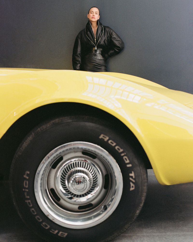 Fashion shot of an adult woman in a black leather outfit, leaning on the hood of a bright yellow vintage car in a studio setting.