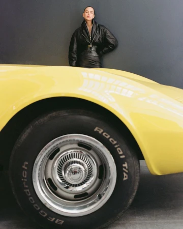 Fashion shot of an adult woman in a black leather outfit, leaning on the hood of a bright yellow vintage car in a studio setting.