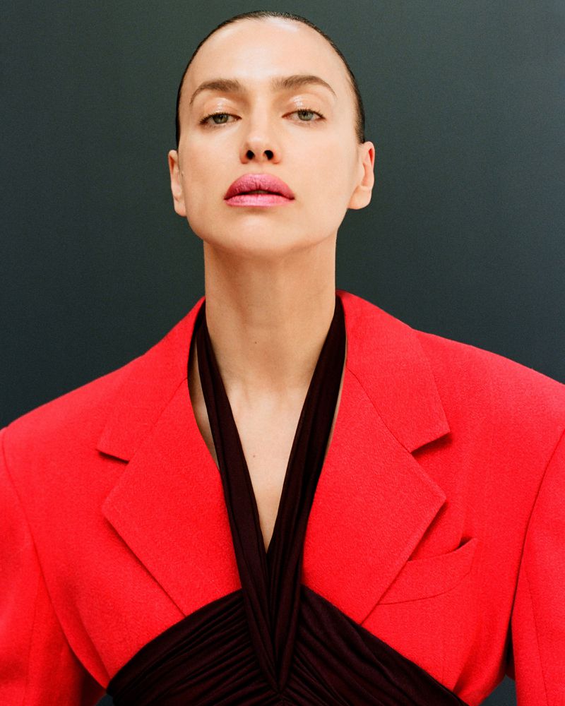 Close-up portrait of a woman with slicked-back hair wearing a red jacket with a black, draped top against a dark grey background.