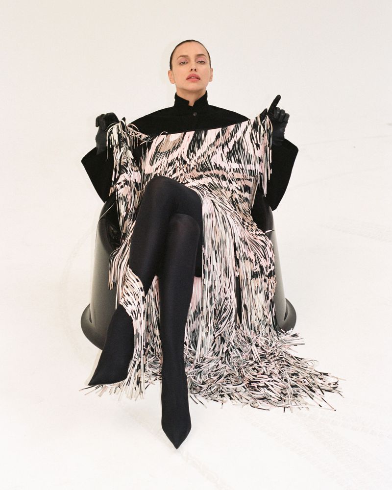 Irina Shayk sits in a black chair wearing black tights and gloves with a black jacket and a dress covered in fringe.