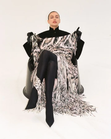 Irina Shayk sits in a black chair wearing black tights and gloves with a black jacket and a dress covered in fringe.