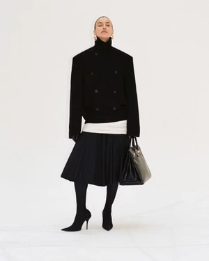 Irina Shayk posing in a high-neck black jacket, pleated skirt, black tights, and pointed boots against a white background.