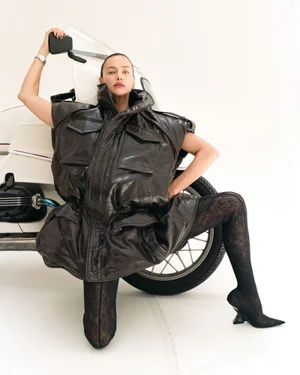 Irina Shayk wearing an oversized dark brown leather puffer vest and lace tights while posing in front of a white motorcycle.