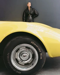 Model Irina Shayk wearing a black leather jacket leaning against a dark wall behind a bright yellow car