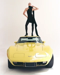Model Irina Shayk standing atop the roof of a vintage yellow sports car in a black mini dress against a white studio background.