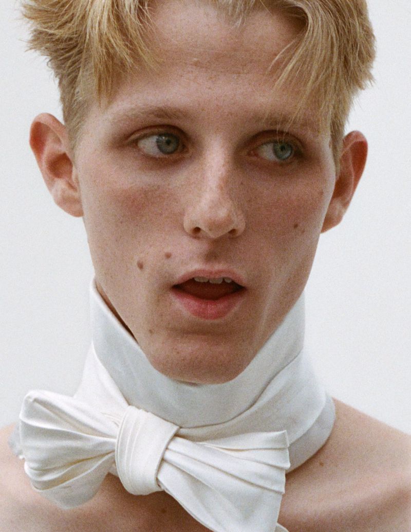 Close-up portrait of a young adult male with blond hair and blue eyes wearing a white bow tie against a bright neutral background.