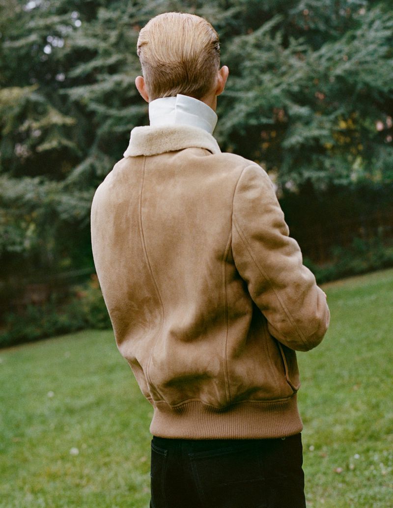 Back view of a young adult in a tan jacket and white turtleneck standing in a grassy, outdoor setting with green trees in the background.