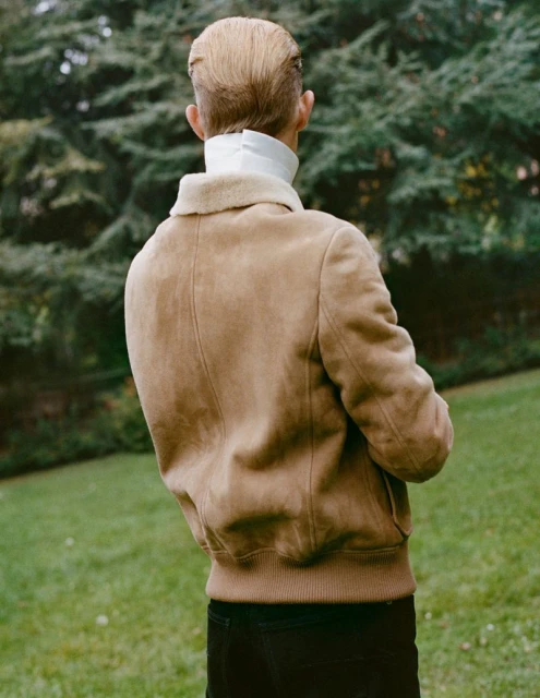 Back view of a young adult in a tan jacket and white turtleneck standing in a grassy, outdoor setting with green trees in the background.
